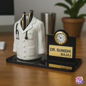 Op de afbeelding: Een witte keramische pennenhouder in de vorm van een doktersjas met stethoscoopdetail, gecombineerd met een zwarte bureauklok met een goudkleurige wijzerplaat en een gouden plaatje met de tekst DR. SUNIDHI BAJAJ op een zwarte rechthoekige voet.