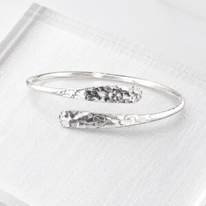 May include: A silver bypass bangle bracelet featuring a polished finish and two tapered, hammered ends. The metal surface reflects light, highlighting the textured, dimpled pattern on the overlapping terminals of this open-style cuff.