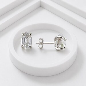 May include: A pair of stud earrings featuring raw, clear, hexagonal-shaped crystals set in silver-toned metal prongs. One earring is shown from the front, while the other is positioned to display the silver post and butterfly clutch backing.