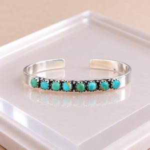 May include: A silver cuff bracelet featuring a row of eight round, polished turquoise stones set in scalloped bezels. The metal band has a smooth, reflective finish and an open back design, showcasing the vibrant blue-green colour of the stones.