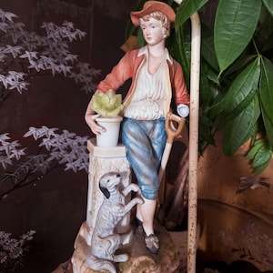 May include: A porcelain figurine of a person wearing a rust-colored jacket, white shirt, and blue trousers, standing next to a white pedestal. The person holds a potted plant and a shovel. A small gray and white dog stands at the base of the pedestal.