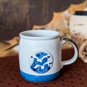 May include: A speckled white ceramic mug featuring a blue band at the base and a circular relief emblem on the front. The emblem displays a blue and white design of two stylized birds in flight. The mug has a curved handle with a dark accent.