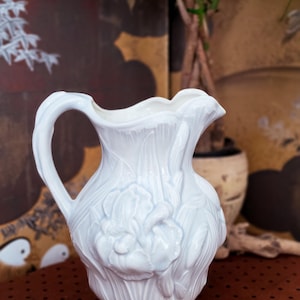 May include: A white ceramic pitcher featuring a raised, embossed floral design of iris flowers and leaves. The pitcher has a curved handle, a scalloped rim, and a glossy finish that reflects light across its textured, sculpted surface.