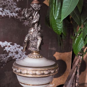 May include: A table lamp featuring a silver-toned metal cherub figure as the central stem. The base is a frosted glass urn-shaped vessel with gold-colored horizontal bands and intricate floral detailing around the middle section.