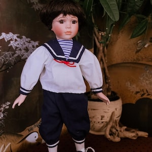 Può includere: Una bambola in porcellana con capelli e occhi castani, che indossa una camicia bianca a maniche lunghe con colletto alla marinara blu navy, una maglietta a righe blu e bianche, lacci rossi, pantaloni blu navy e scarpe stringate bicolore.