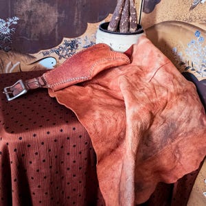 May include: A tan-coloured leather accessory featuring a wide, textured, and irregular-shaped piece of hide attached to a narrow strap with a silver-toned metal buckle. The leather displays visible grain, natural markings, and a stitched edge detail.