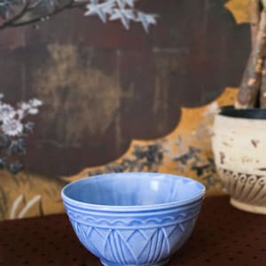 May include: A light blue ceramic bowl featuring a decorative embossed pattern around the exterior. The bowl has a rounded shape with a smooth interior and a textured, geometric design consisting of scalloped arches and vertical lines on the outside.