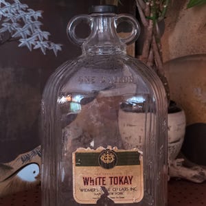 May include: A clear glass one-gallon jug with two circular handles at the top and a black cap. The bottle features vertical ribbed detailing and a rectangular paper label that reads White Tokay, Widmer's Wine Cellars Inc, Naples, New York.