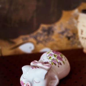 May include: A white ceramic pig figurine in a reclining position with closed eyes. The pig features a painted pink snout, a small purple flower on its cheek, and a light purple bow between its ears. A cluster of purple and pink floral patterns adorns its back.