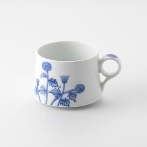 以下が含まれることがあります： 外側にアザミの花と葉の青い植物画が描かれた白い陶磁器のカップ。カップは幅広で背の低い円筒形をしており、上部に小さな青い花の模様があしらわれた円形のハンドルが付いています。