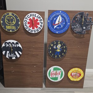 Könnte beinhalten: Acht runde Wanduhren auf zwei braunen Holzplatten. Die Designs zeigen Nirvana, Red Hot Chili Peppers, Starfleet Command, Star Wars, The Beatles, eine Fallout-Figur sowie Castrol- und Shell-Logos, jeweils mit goldfarbenen Uhrzeigern.