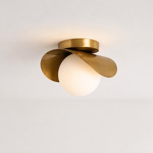 May include: A flush mount ceiling light fitting featuring a central spherical white glass globe surrounded by a curved, petal-like brass shade. The metallic brass component has a smooth, reflective finish, while the globe emits a soft glow.