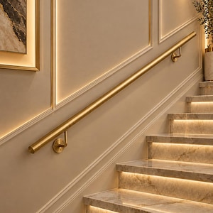 Op de afbeelding: Een handgreep van geborsteld goudkleurig metaal gemonteerd op een beige muur met decoratieve lijsten. De trap heeft marmeren treden met geïntegreerde warme LED-stripverlichting die de randen van elke trede langs de muur verlicht.