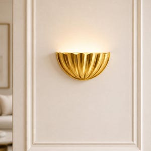 Könnte beinhalten: Eine halbkreisförmige Wandleuchte mit goldener Metalloberfläche und einem gewellten, muschelartigen Design. Die Leuchte ist an einer flachen Oberfläche montiert und strahlt ein warmes, sanftes Licht aus ihrer oberen Öffnung aus.