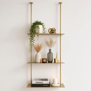 May include: A three-tier gold metal wall-mounted shelving unit with two vertical support poles. The shelves hold a trailing green plant in a white pot, a gold knot sculpture, dried grasses in vases, a candle, and books labeled MINIMAL LIVING.