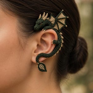 May include: A dark green and tan crocheted dragon ear cuff. The dragon features a detailed head with small horns, textured wings, and a long, winding body that curves around the outer edge of the ear, ending in a pointed, leaf-shaped tail.