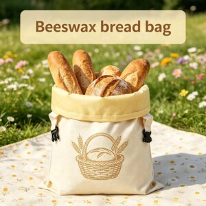 May include: A cream-colored beeswax bread bag with a yellow interior lining and black side buckles. The front features a brown graphic of a loaf of bread in a basket with wheat stalks. The text "Beeswax bread bag" is printed at the top.