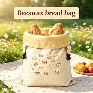 May include: A cream-colored beeswax bread bag featuring a printed pattern of various pastries and small stars. The bag has a wide, folded-over top edge and black plastic side buckles. Two loaves of crusty bread are visible inside the open bag.