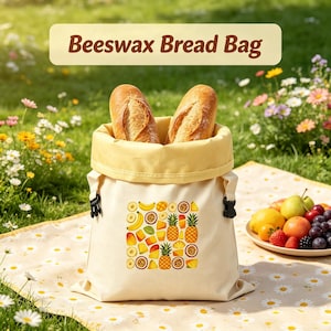 Puede incluir: Una bolsa de cera de abeja para pan de color crema con un estampado gráfico cuadrado de varias frutas tropicales, incluyendo piñas, plátanos y maracuyá. La bolsa tiene un forro interior amarillo claro, hebillas laterales negras y dos barras de pan.