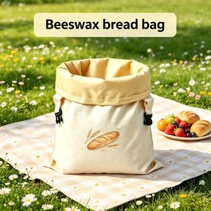 Puede incluir: Una bolsa para pan de cera de abeja de color crema con un borde superior enrollado y hebillas laterales de plástico negro. El frente presenta una ilustración gráfica de una hogaza de pan con espigas de trigo y el texto Beeswax bread bag.