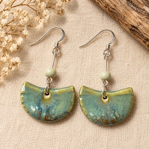 May include: A pair of dangle earrings featuring semi-circular ceramic pendants with a glossy teal and yellow-green glaze. Each pendant hangs from a silver-toned metal hook and is accented with a single small, pale green spherical bead.