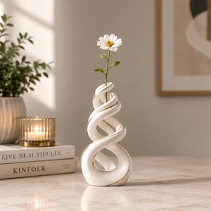 May include: A white ceramic vase with a spiraling, twisted design and gold-toned trim along the edges. The vase features a single opening at the top, currently holding a white daisy with a yellow center. The structure is hollow and sculptural.