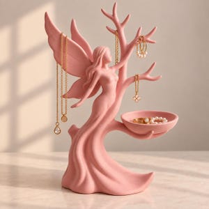 May include: A matte pink jewellery organiser shaped like a fairy with wings standing next to a tree. The tree branches hold gold necklaces and hoop earrings, while a small attached bowl holds rings and a pearl bracelet. The figure has a smooth finish.