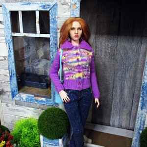May include: A doll wearing a purple and pink patterned cardigan with a button closure, blue jeans, and light blue shoes. The doll has long, red hair and is standing in front of a wooden door with a blue frame.