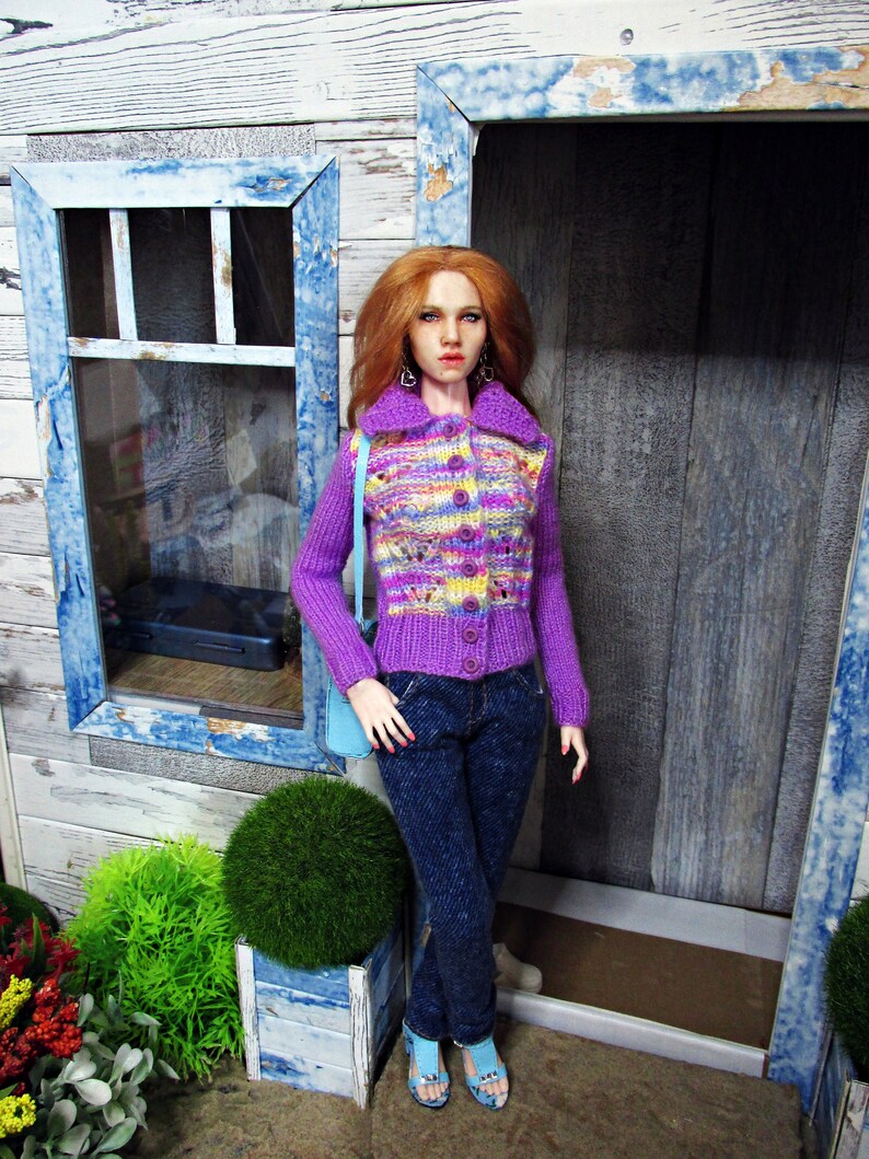 May include: A doll wearing a purple and pink patterned cardigan with a button closure, blue jeans, and light blue shoes. The doll has long, red hair and is standing in front of a wooden door with a blue frame.