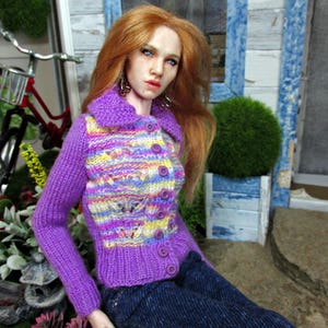 May include: A doll wearing a purple and yellow striped knitted cardigan with a button closure. The doll has long red hair and blue eyes. The doll is wearing blue jeans and is sitting on a green surface.