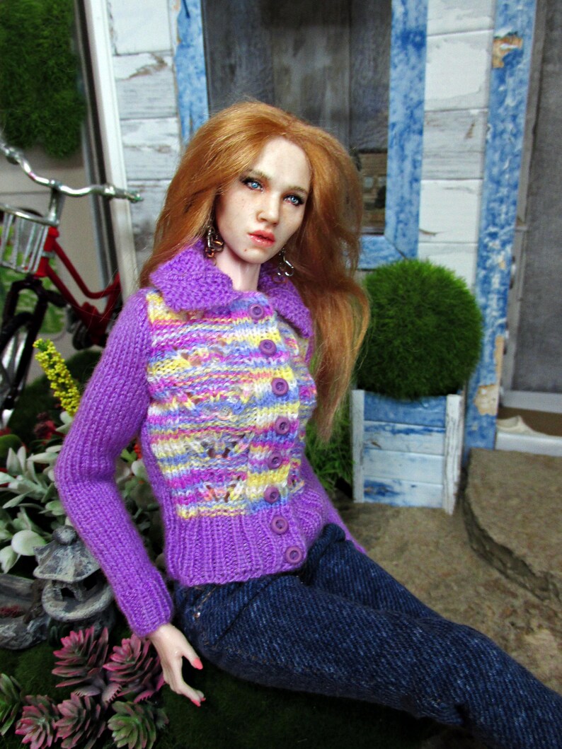 May include: A doll wearing a purple and yellow striped knitted cardigan with a button closure. The doll has long red hair and blue eyes. The doll is wearing blue jeans and is sitting on a green surface.