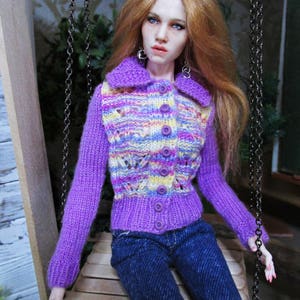 May include: A doll wearing a purple, yellow, and pink striped cardigan with a button closure and blue jeans.