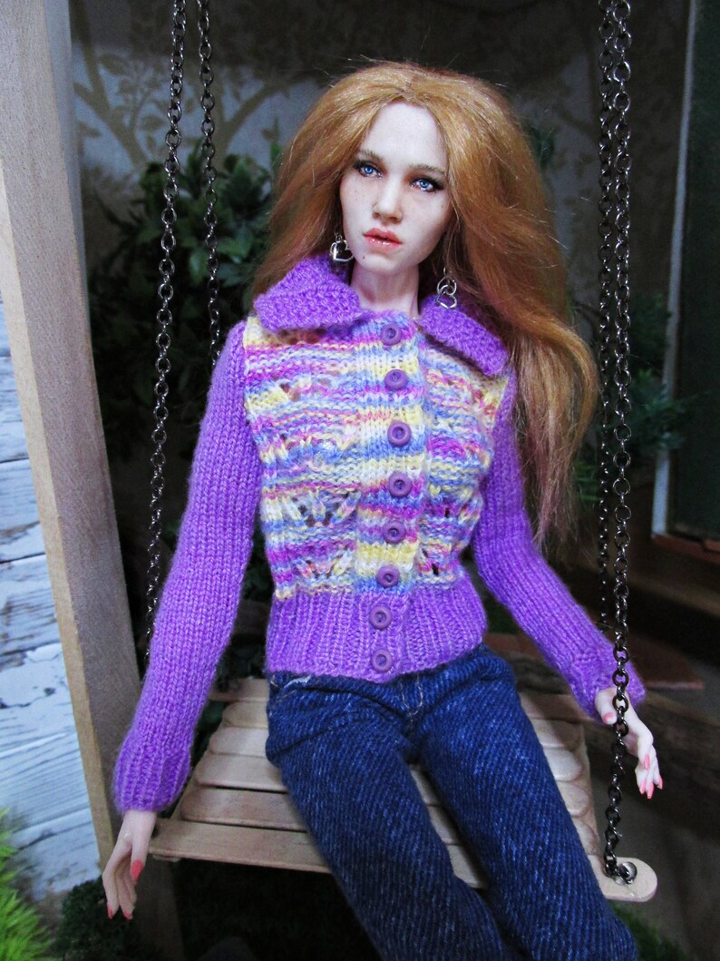May include: A doll wearing a purple, yellow, and pink striped cardigan with a button closure and blue jeans.