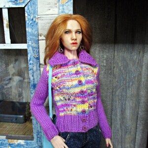 May include: A doll wearing a purple and multi-colored knitted cardigan with a light blue shoulder bag. The cardigan has a button closure and a cable knit pattern. The doll has long red hair and is wearing blue jeans.