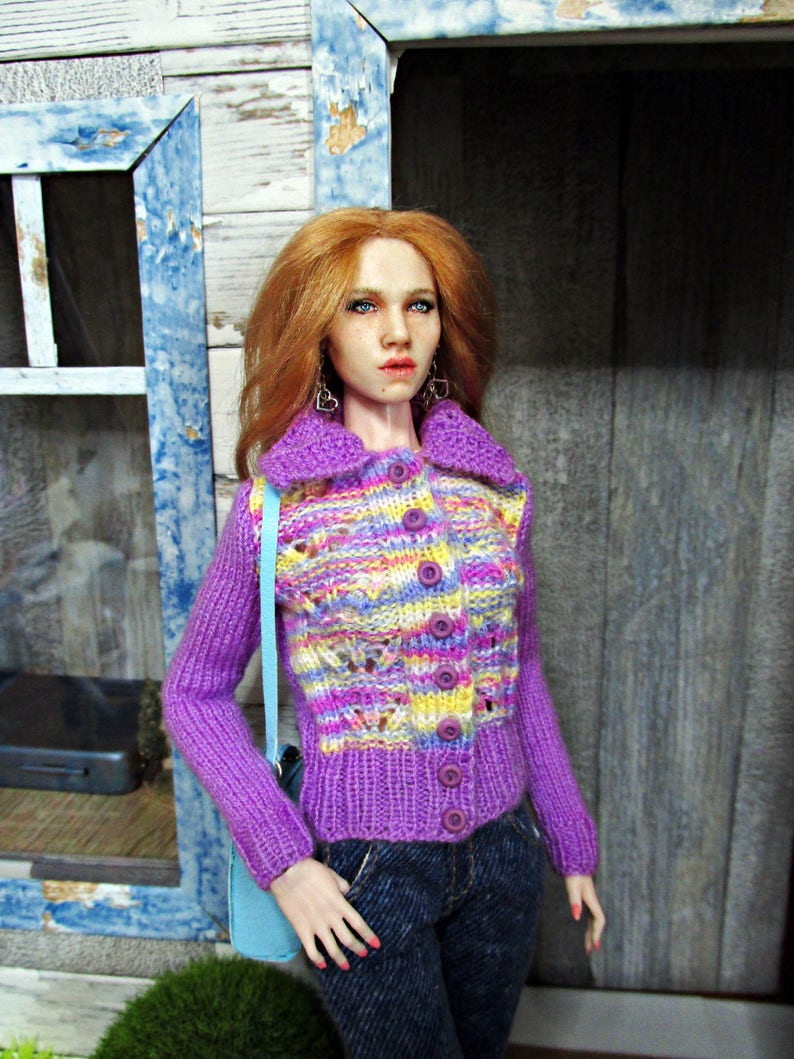 May include: A doll wearing a purple and multi-colored knitted cardigan with a light blue shoulder bag. The cardigan has a button closure and a cable knit pattern. The doll has long red hair and is wearing blue jeans.