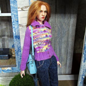May include: A doll wearing a purple and multi-colored knitted cardigan with buttons, blue jeans, and a light blue handbag.
