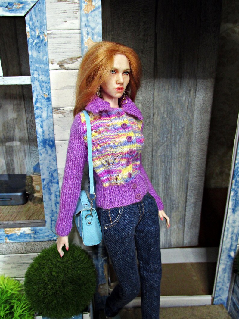 May include: A doll wearing a purple and multi-colored knitted cardigan with buttons, blue jeans, and a light blue handbag.