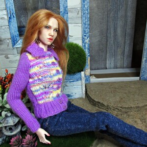 May include: A doll wearing a purple and yellow striped knitted cardigan with a blue denim skirt. The doll has long red hair and is sitting on a wooden surface.