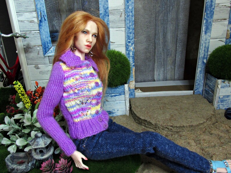 May include: A doll wearing a purple and yellow striped knitted cardigan with a blue denim skirt. The doll has long red hair and is sitting on a wooden surface.