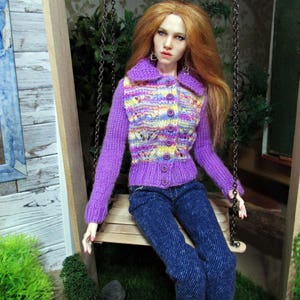 May include: A doll wearing a purple and yellow striped sweater with a button closure, blue jeans, and red nail polish. The doll is sitting on a wooden swing.