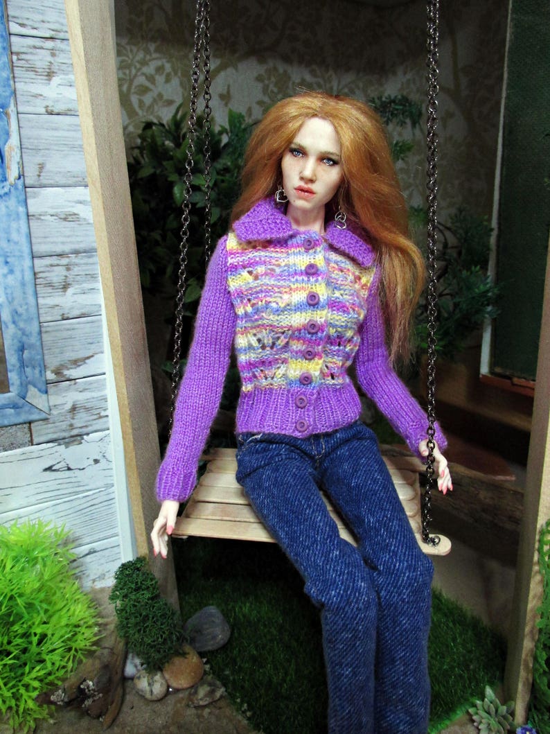 May include: A doll wearing a purple and yellow striped sweater with a button closure, blue jeans, and red nail polish. The doll is sitting on a wooden swing.