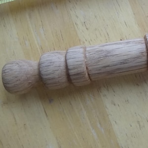 Puede incluir: Un rodillo de madera con un acabado marrón claro. El rodillo tiene un cuerpo cilíndrico con una serie de anillos decorativos y un mango redondeado en un extremo. El otro extremo tiene un mango más pequeño y redondeado.