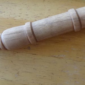 Puede incluir: Un rodillo de madera de color claro con un mango redondeado y dos anillos decorativos. El rodillo es de forma cilíndrica y parece estar hecho de madera sin terminar. El mango es bulboso y tiene una pequeña hendidura en el extremo.