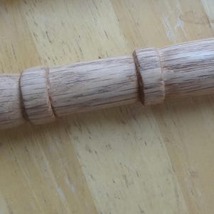 Puede incluir: Un rodillo de madera con un acabado marrón claro. El rodillo tiene un cuerpo cilíndrico con un mango redondeado en un extremo y un mango decorativo más pequeño en el otro. La veta de la madera es visible.