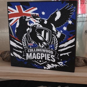 May include: A square black plaque featuring a graphic of three magpies, the Australian flag, and a shield emblem. The text "COLLINGWOOD MAGPIES" is printed in white capital letters across the center. The design has a textured, glittery finish.