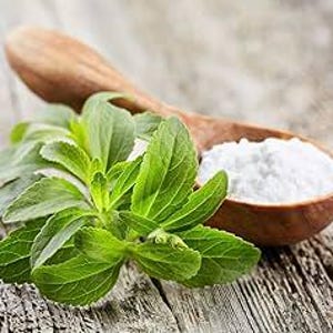 May include: A close-up shot shows fresh green stevia leaves next to a wooden spoon filled with white powder, likely stevia extract. The spoon and leaves rest on a weathered wooden surface, suggesting a natural, organic theme.