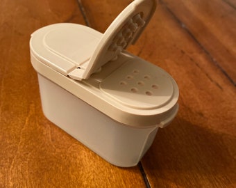 May include: A white plastic spice shaker with a flip-top lid and a small hole for dispensing spices.