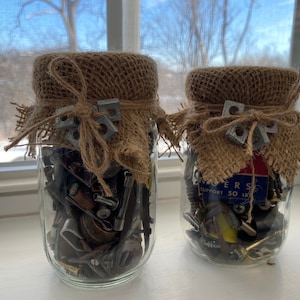 May include: Two glass jars filled with assorted hardware and metal pieces, covered with burlap fabric and tied with twine. The jars are sitting on a white surface.