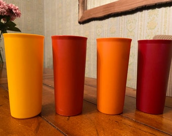May include: Four tall, cylindrical drinking glasses in different shades of orange, red, and yellow. The glasses are arranged in a row from left to right, with the yellow glass on the left, followed by the orange, red, and then the darkest red glass on the right.