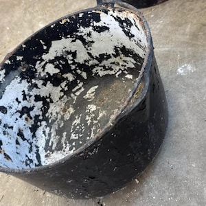 May include: A round, deep metal pot with a black exterior and a distressed white interior finish. The vessel features a wide opening, a curved rim, and small metal loops on opposite sides for a handle attachment, showing signs of wear.